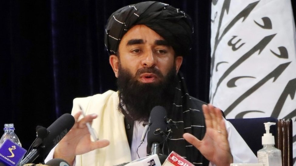 Taliban officials speaking at press conference with Afghan flags in background