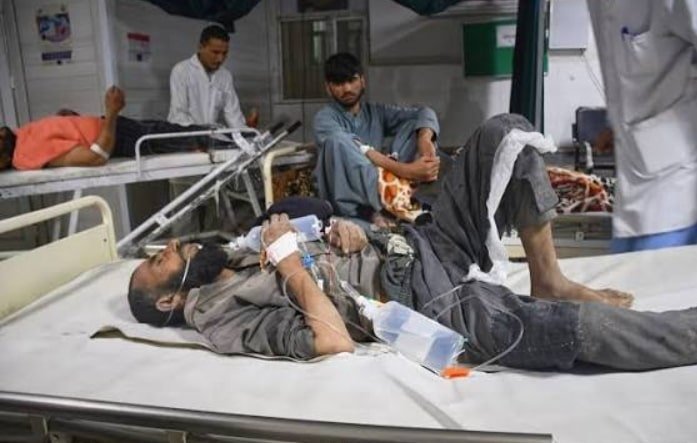 A man lies on a hospital bed in a crowded ward, his legs extended and watching staff attend to him, while other injured patients and medical workers surround him in the background.